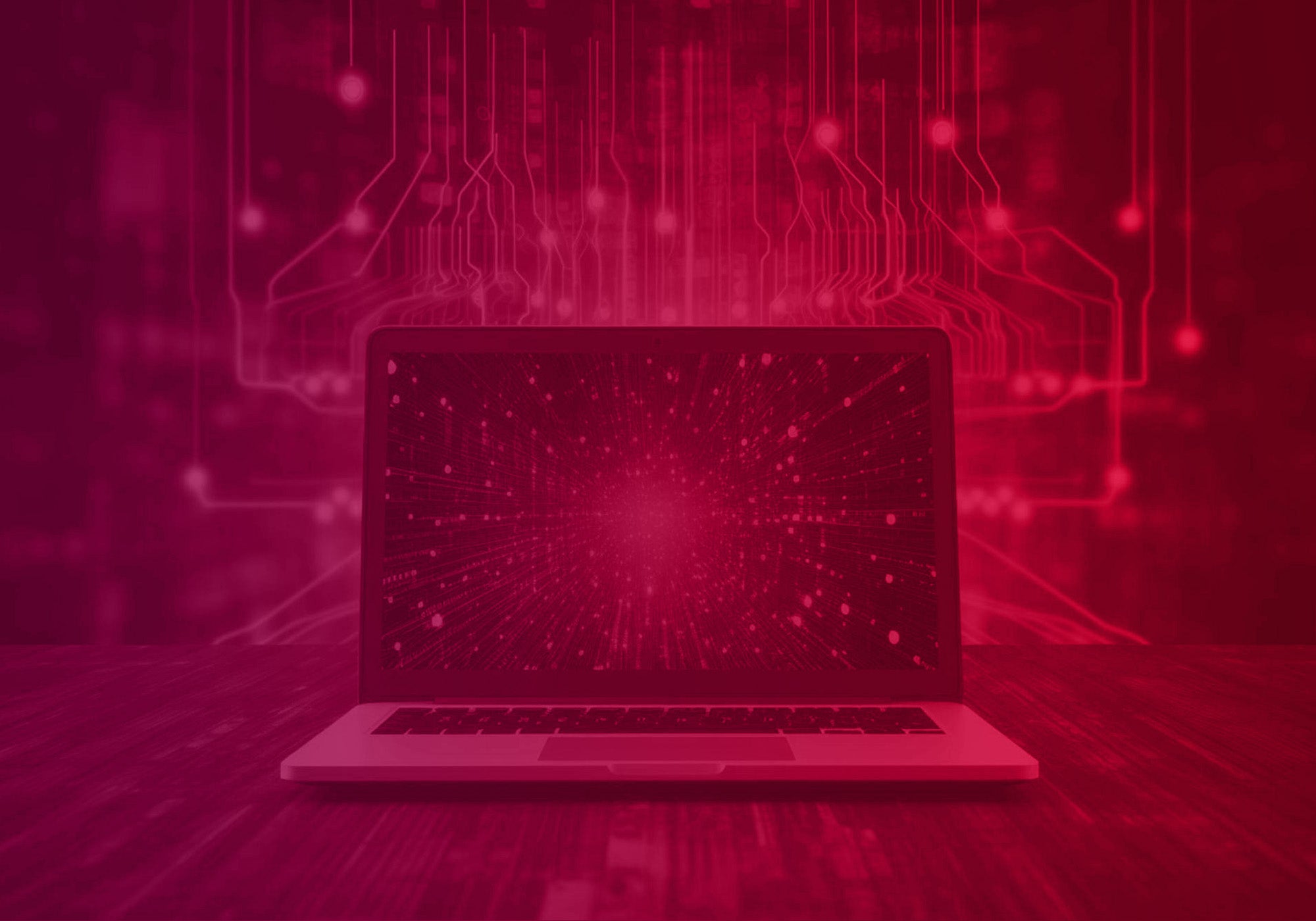 An open laptop sits on a wooden surface, with a glowing digital network or circuit-board graphic in red and magenta tones behind it.