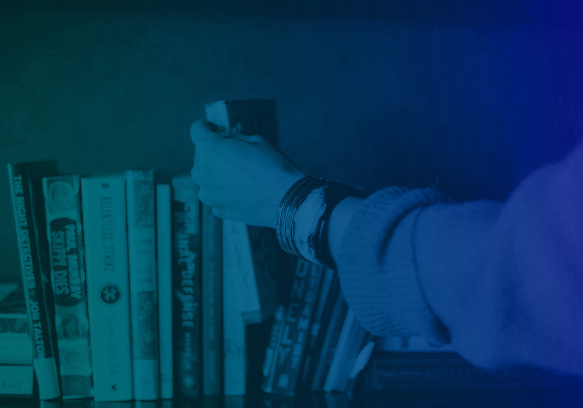 A person's hand and arm, wearing a light-colored knit sweater and bracelets, reaching to adjust a row of books on a shelf under a strong blue and teal tint.