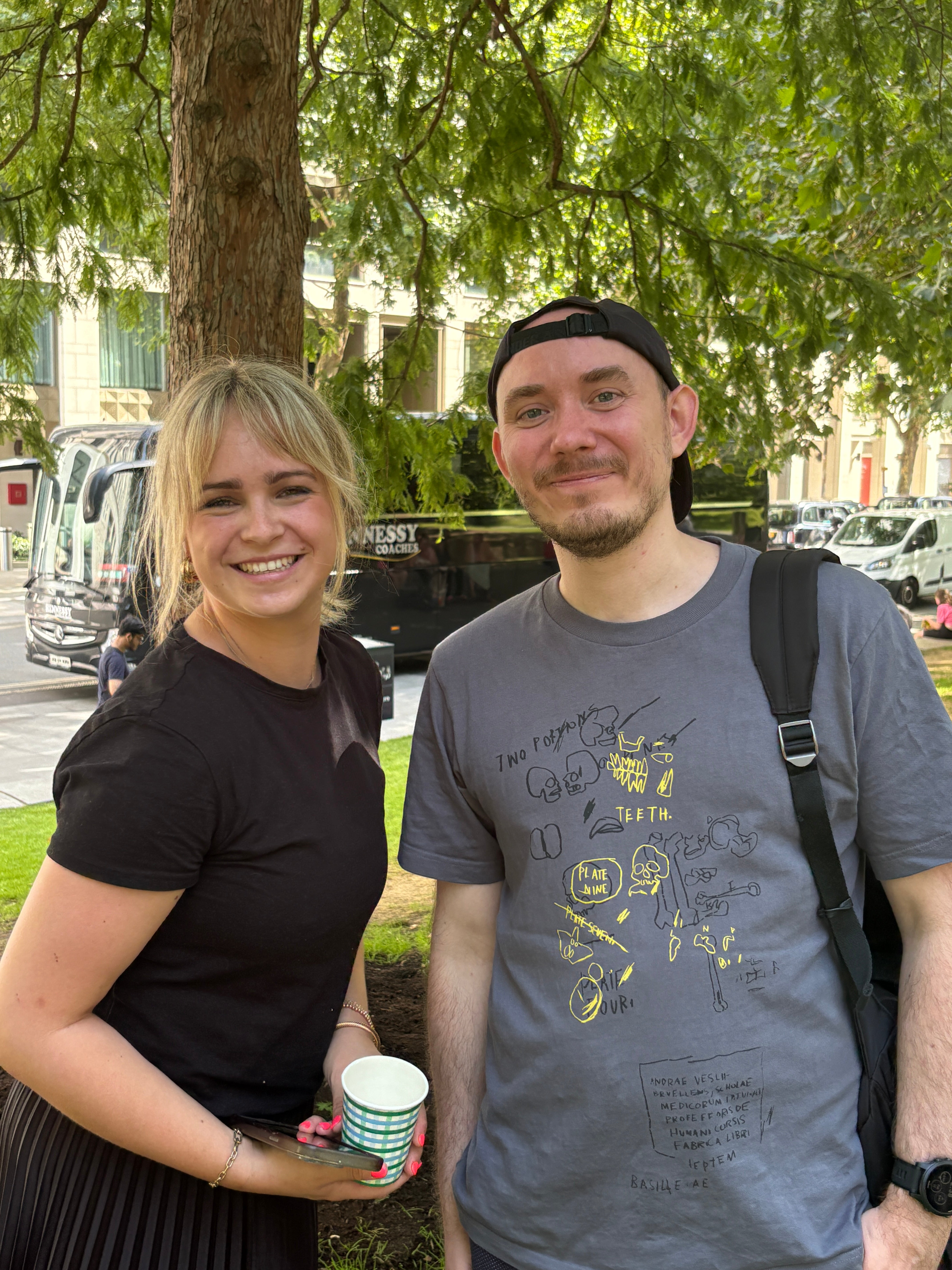 Two people standing outdoors with trees and a street in the background