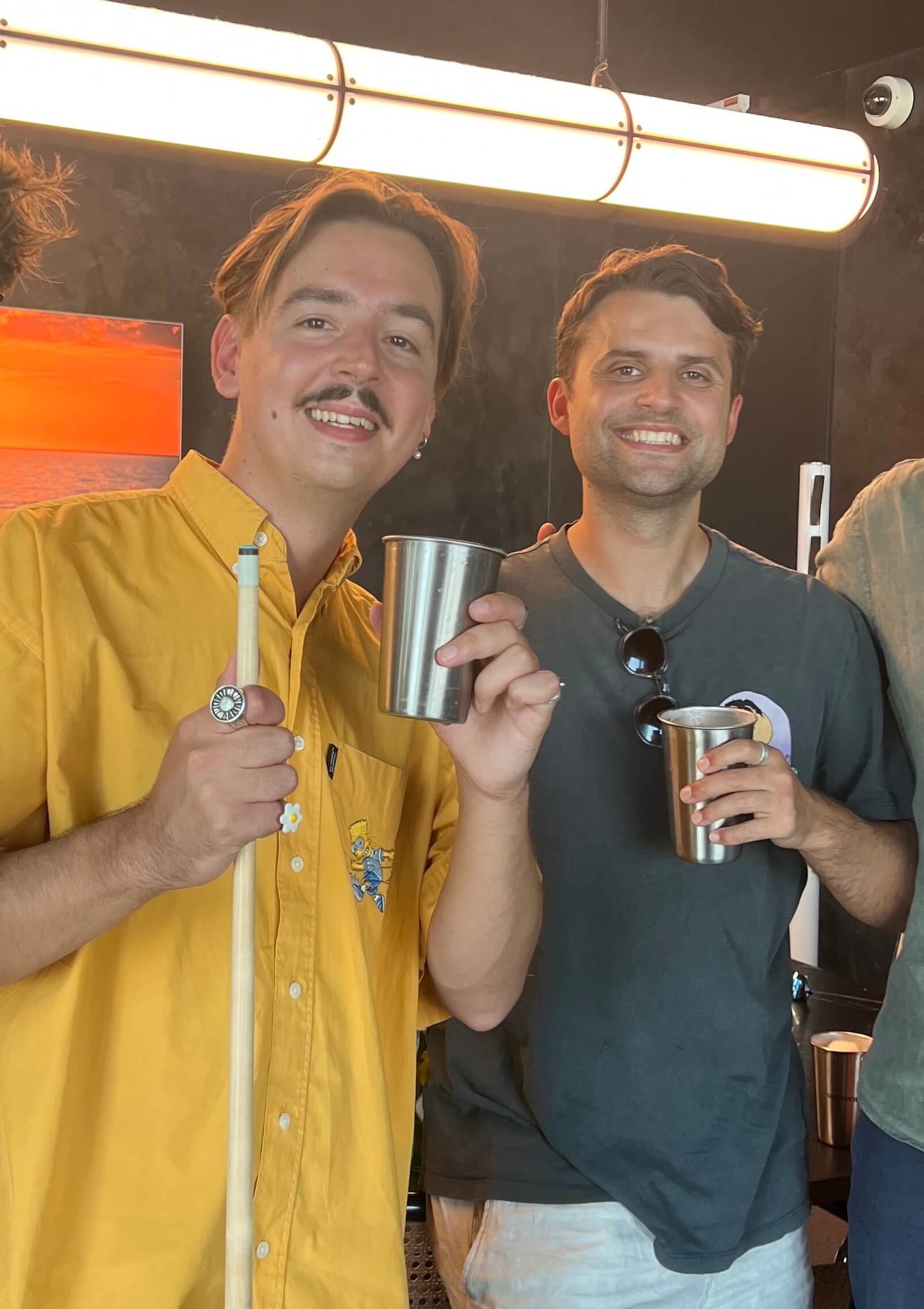 Two men holding metal cups and a pool cue in a casual setting.