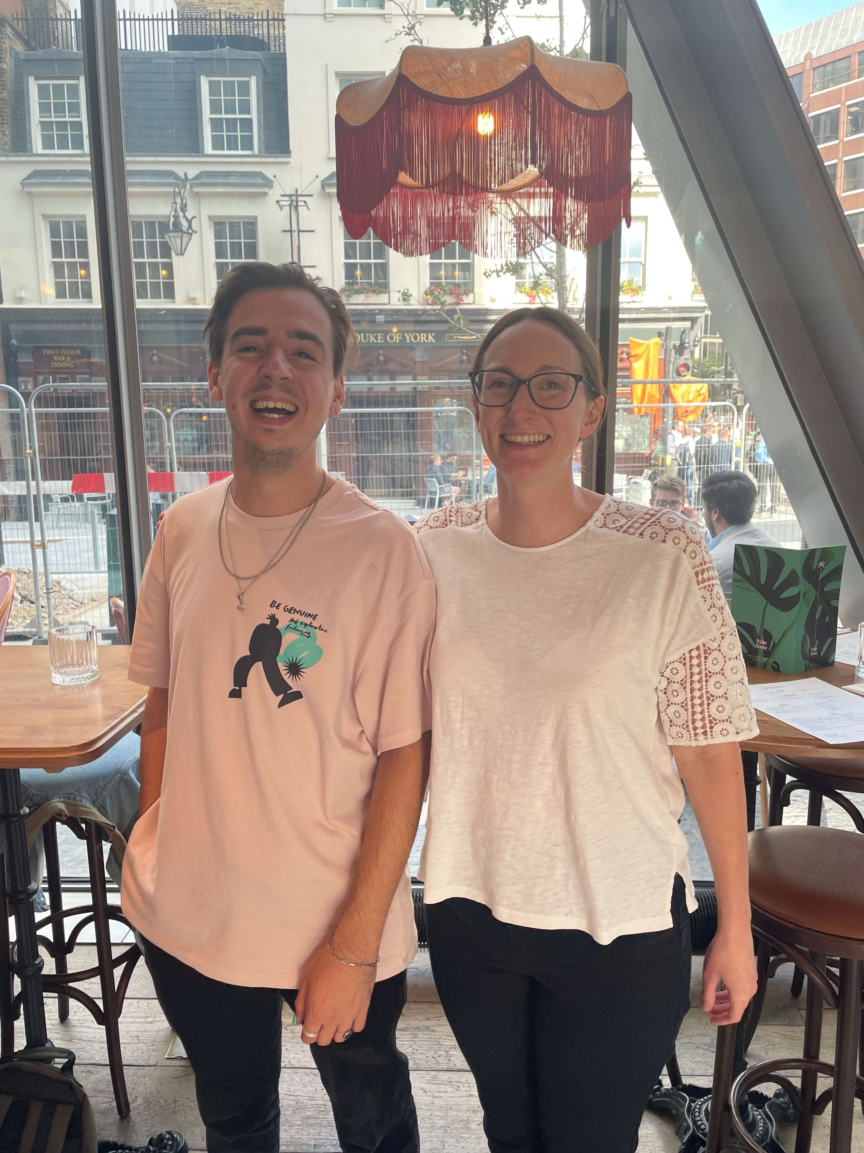 Two people standing in a casual setting with a window view of an urban street.