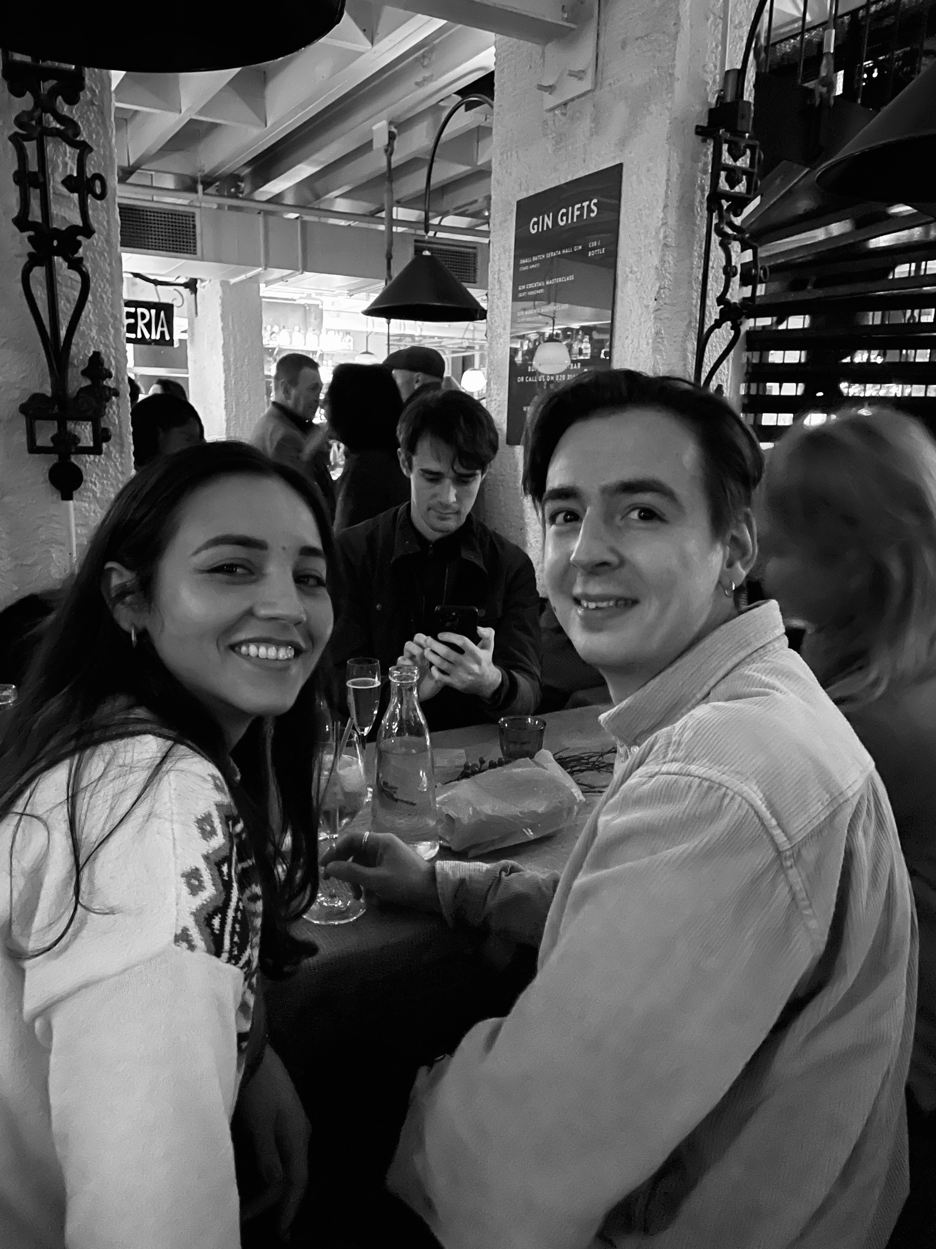 Two people sitting together in a social setting with other people in the background.