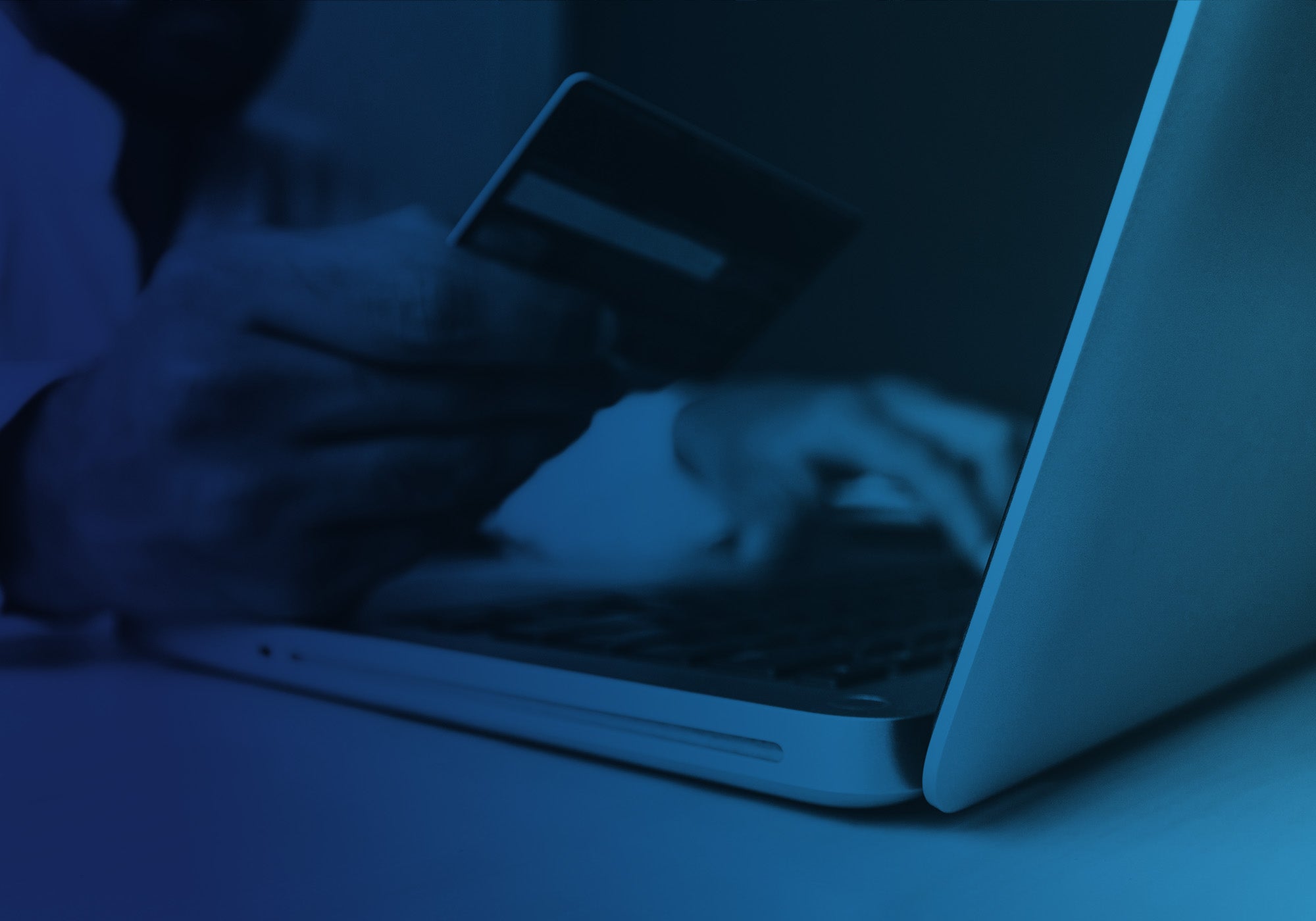 A person using a laptop and holding a credit card, shown in a heavily blue-toned, moody photograph.