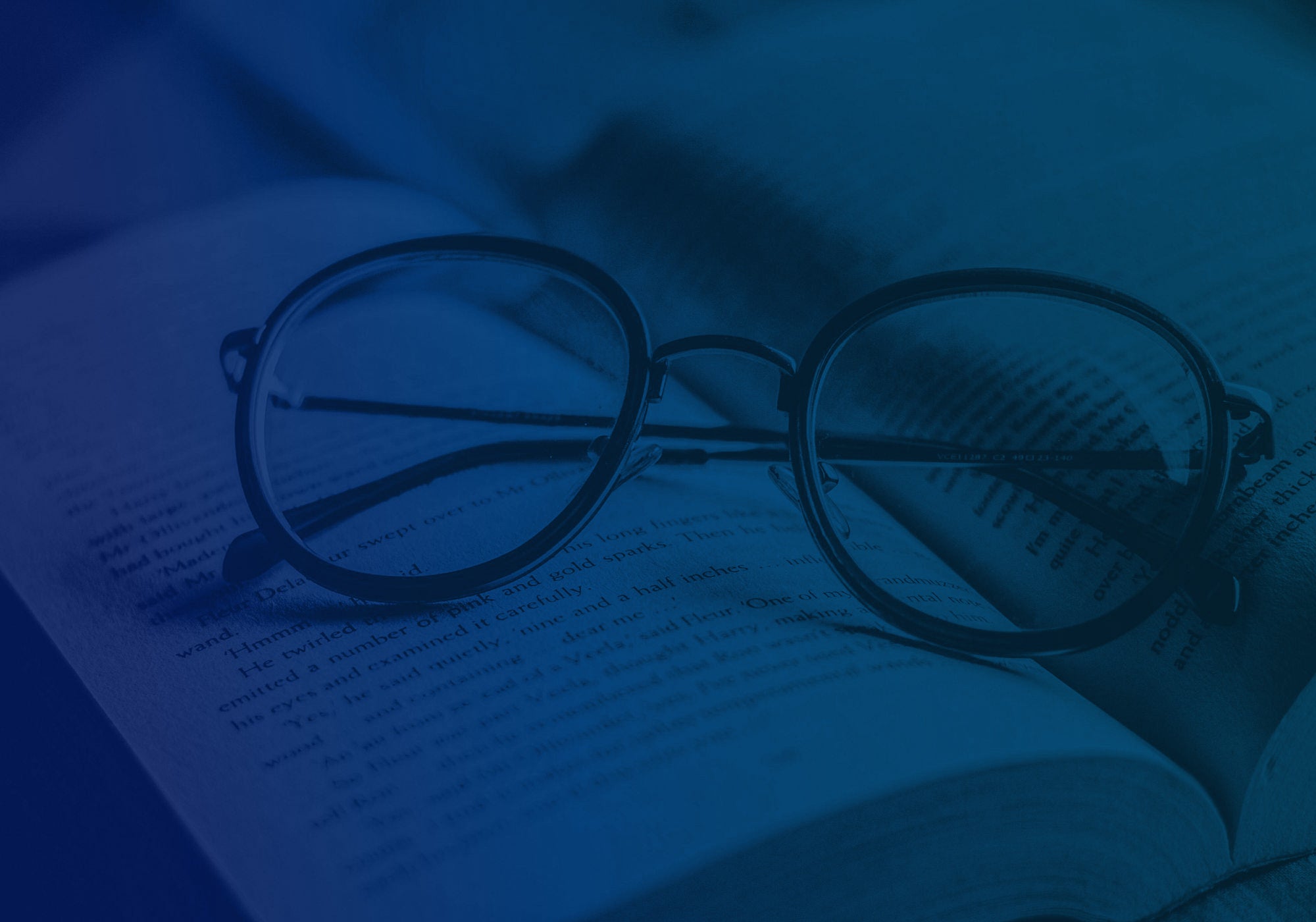 Round metal eyeglasses resting on an open book, shown in a blue-tinted close-up shot.